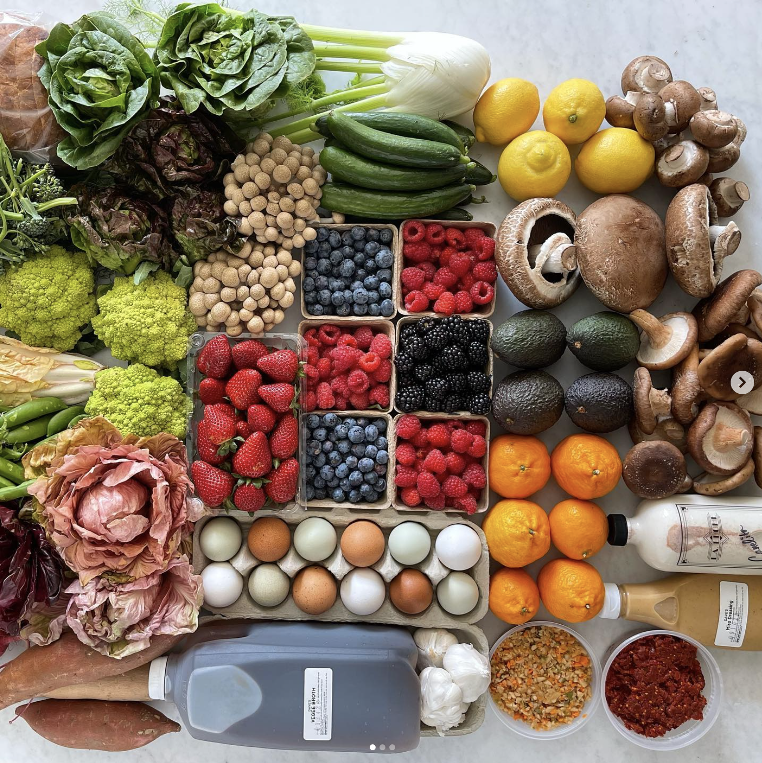 Colorful array of fruits and vegetables displayed on a table, including eggs, oranges, avocado, assorted berries, cucumbers, lemons, mushgrooms, lettuces