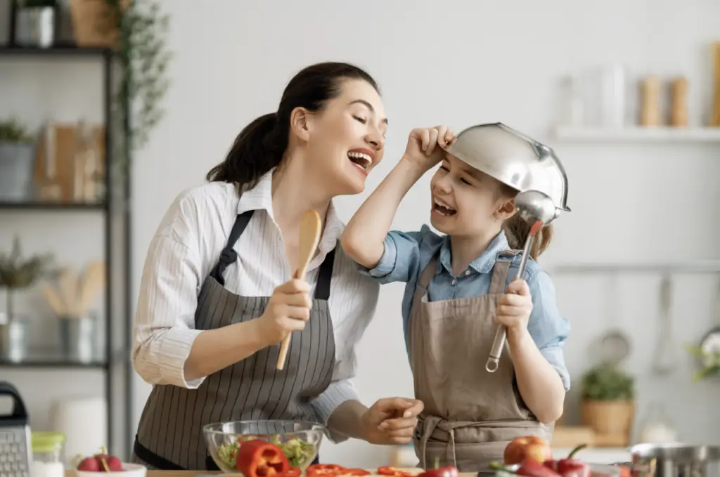 A white woman smiles holding a wooden spoon, while her daughter laughs with a metal mixing bowl on top of her head, standing in the kitchen