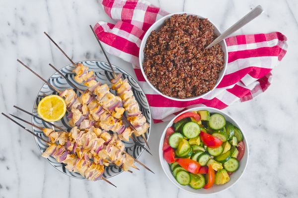 6 skewers of chicken kabobs on a marble table top next to a bowl of quinoa and a bowl of sliced cucumber and tomato salad