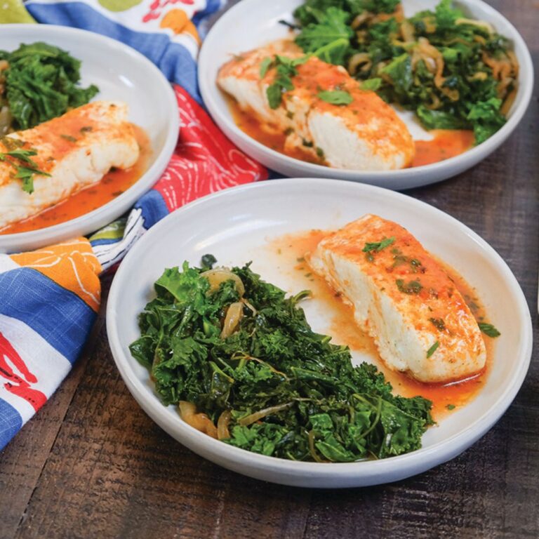 3 plates of sea bass filets with a topping of light oven-roasted blistered grape tomato sauce on top, on a white plate with a side of sautéed kale and sweet onions